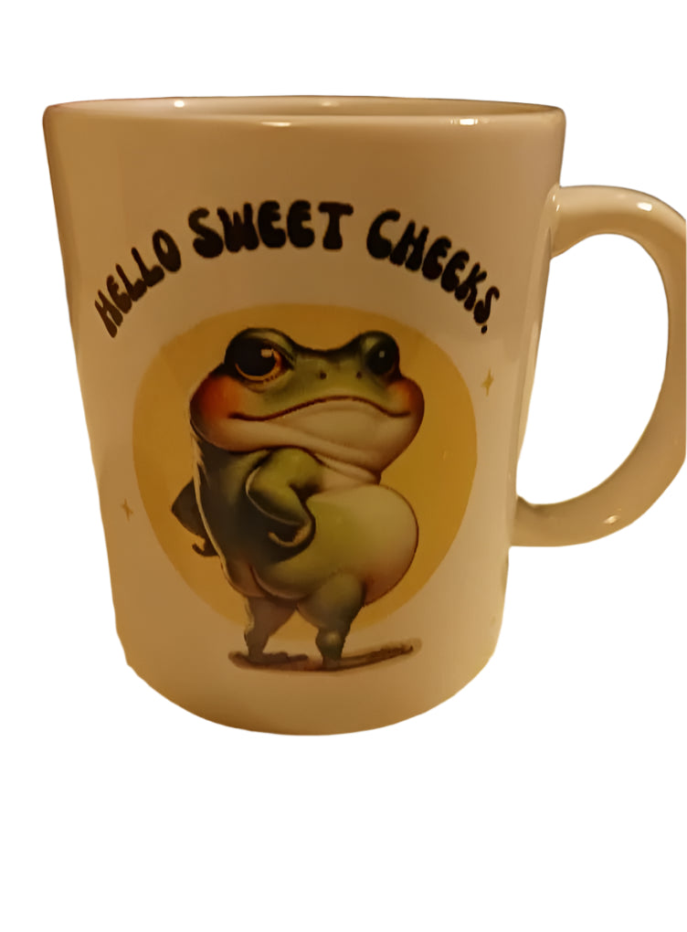 Mug with a frog illustration and 'Hello Sweet Cheeks' text on a yellow patterned surface.